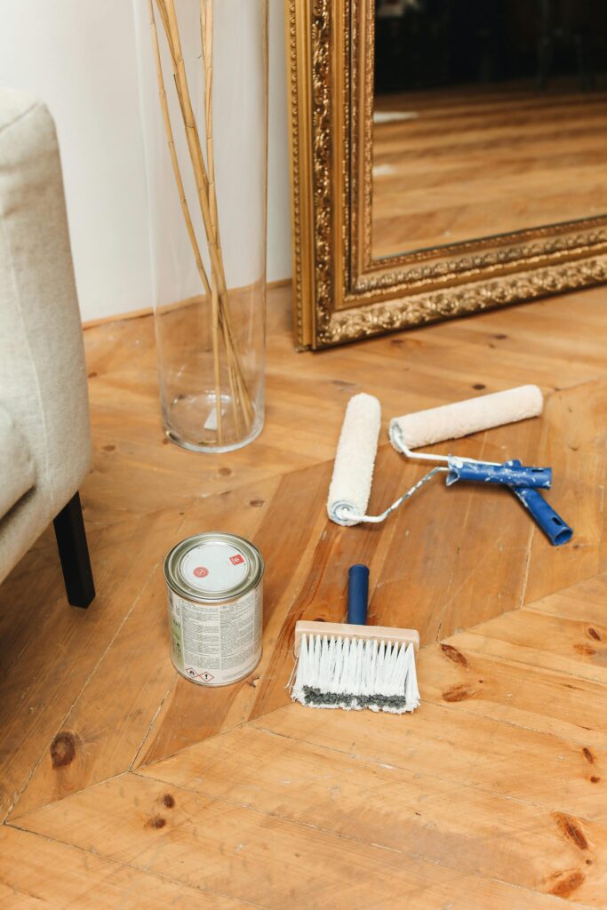 Paint rollers, brush, and paint can on wooden floor for DIY home renovation.
